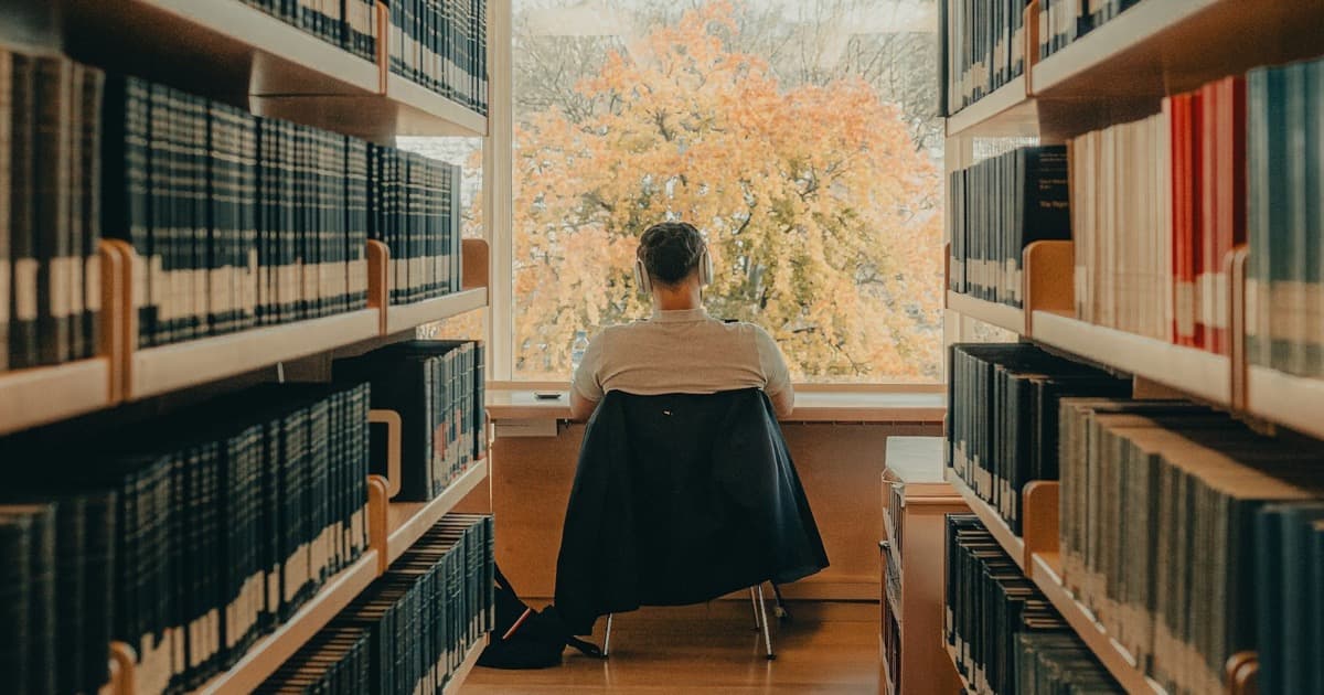 Person mit Kopfhörern sitzt am Fenster am Ende eines Bibliotheksgangs